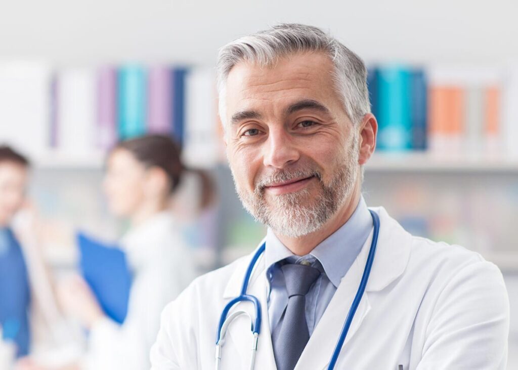 An older doctor looking at the camera with colleagues in the background.